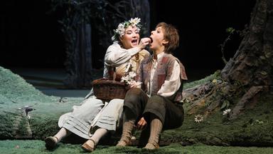 Gretel gibt Hänsel etwas zu essen. Sie sitzen auf einem Waldboden vor Bäumen.