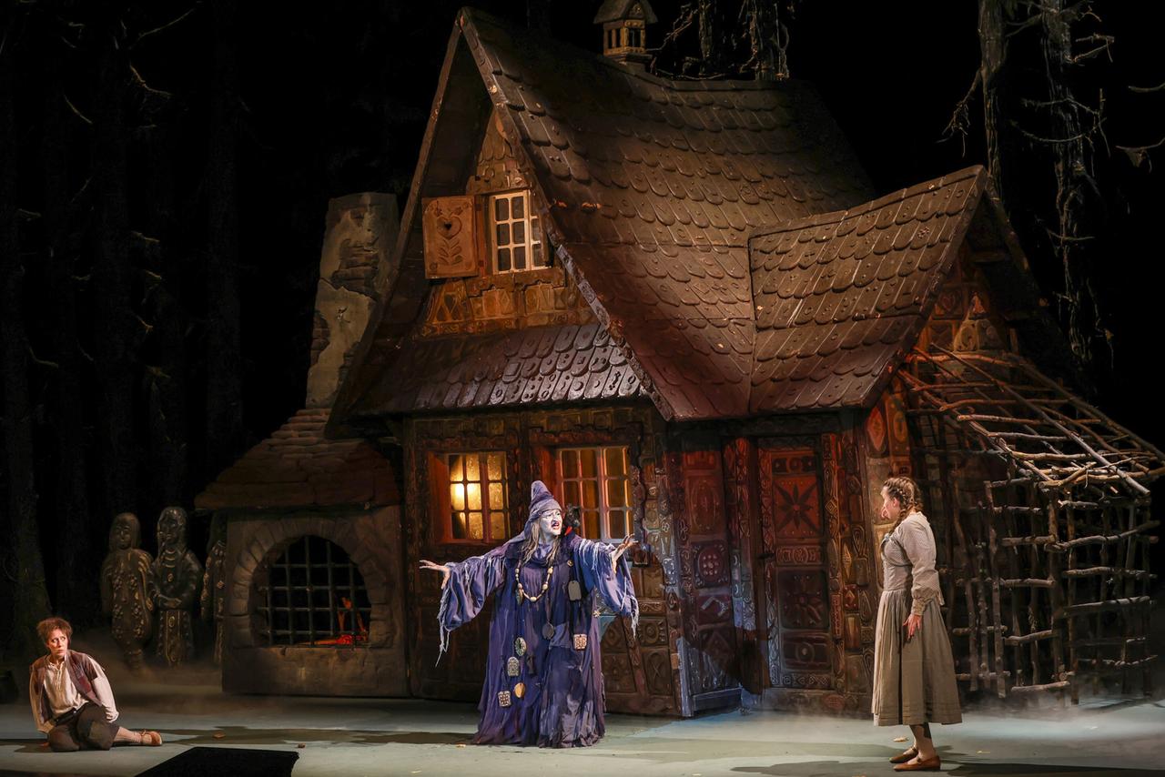 Eine lila gekleidetet Hexe vor ihrem Hexenhaus lockt Hänsel und Gretel herbei