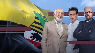 Dieter Hallervorden, Jo Schück und Hans-Tomas Tillschneider stehen nebeneinander und schauen den Betrachter an. Im Hintergrund flattert eine Sachsen-Anhalt-Flagge.