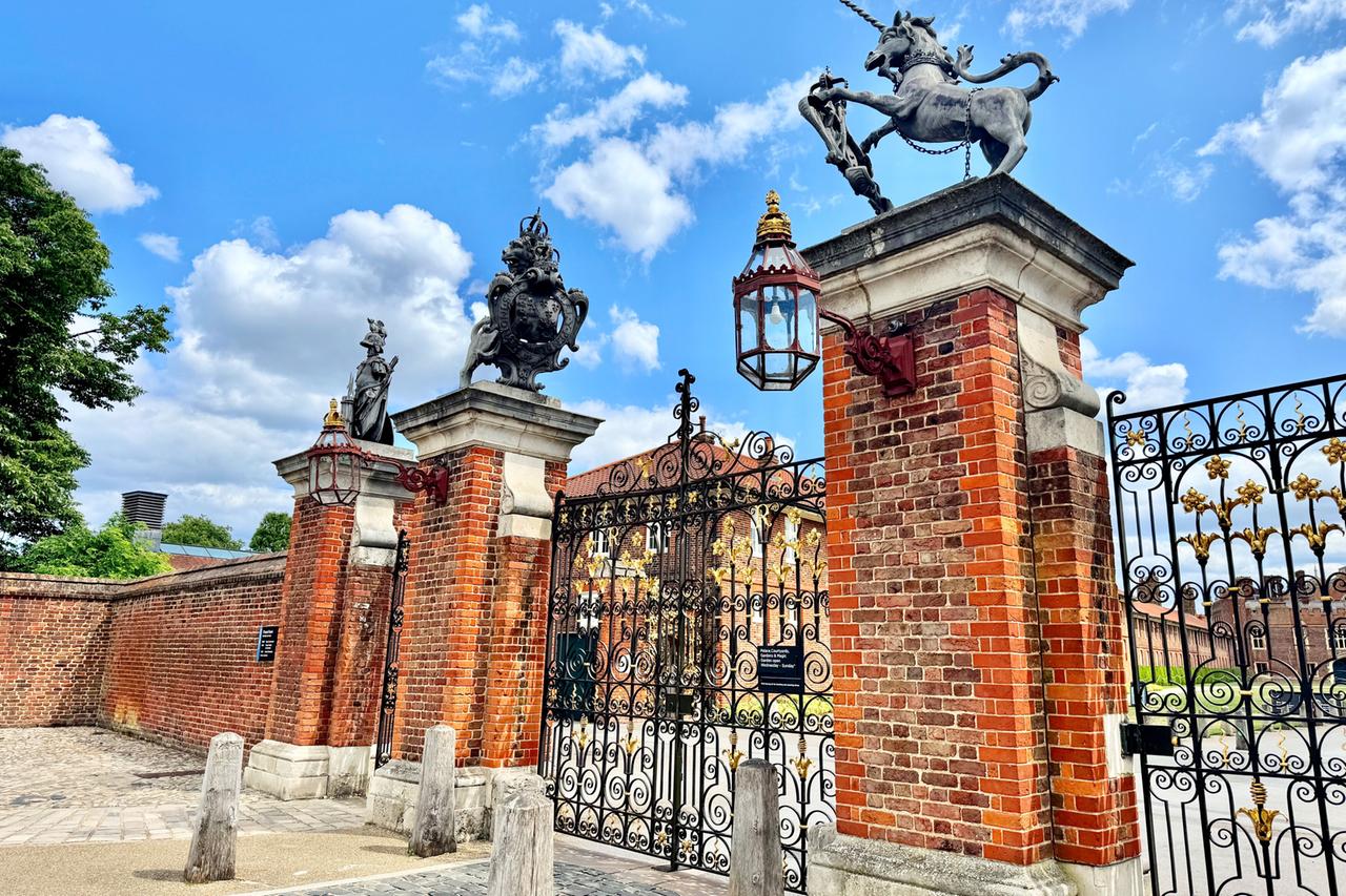 Das Bild zeigt das Haupttor (Main Gate) von Hampton Court Palace, einem historischen Gebäude in England.   Im Vordergrund stehen zwei hohe Pfeiler aus roten Ziegelsteinen, an deren Spitzen sich auf der linken Seite eine Statue einer weiblichen Figur und auf der rechten Seite ein Wappen mit einem steinernen Einhorn befindet. Zwischen den Pfeilern ist ein kunstvoll gestaltetes, schmiedeeisernes Tor sichtbar, das mit goldenen Verzierungen und geschwungenen Mustern versehen ist. An der linken Gebäudeseite hängt eine Leuchte aus Glas und Metall.   Im Hintergrund sind Teile des Palastes und der weitläufige Garten zu erkennen, die von einer Mauer eingerahmt werden. Der Himmel zeigt sich in hellblau mit einigen weißen Wolken, und im Bild ist eine grüne Baumreihe sichtbar. Die Gesamtatmosphäre des Bildes vermittelt den historischen und künstlerischen Wert des Ortes.