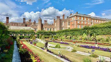 Das Bild zeigt die königlichen Gärten von Hampton Court Palace. Im Vordergrund sind prachtvolle barocke Gärten zu sehen, die mit bunten Blumenbeeten in verschiedenen Formen und Farben gestaltet sind. Ein schmaler Weg aus Stein zieht sich durch die Gartenanlage.   Im Hintergrund erhebt sich das Gebäude des Hampton Court Palace, das aus roten Ziegelsteinen und grauem Stein besteht. Es ist von hohen, grünen Hecken umgeben, die eine klare Abgrenzung zu den Gartenbereichen bilden. Die Fassade des Palastes zeigt verschiedene architektonische Details, darunter runde Fenster und steinerne Verzierungen.   Der Himmel ist blau mit einigen weißen, wolkigen Strukturen, die für eine helle und freundliche Atmosphäre sorgen. Das Bild vermittelt einen Eindruck von harmonischer Gartenkunst, Geschichte und künstlerischem Erbe.