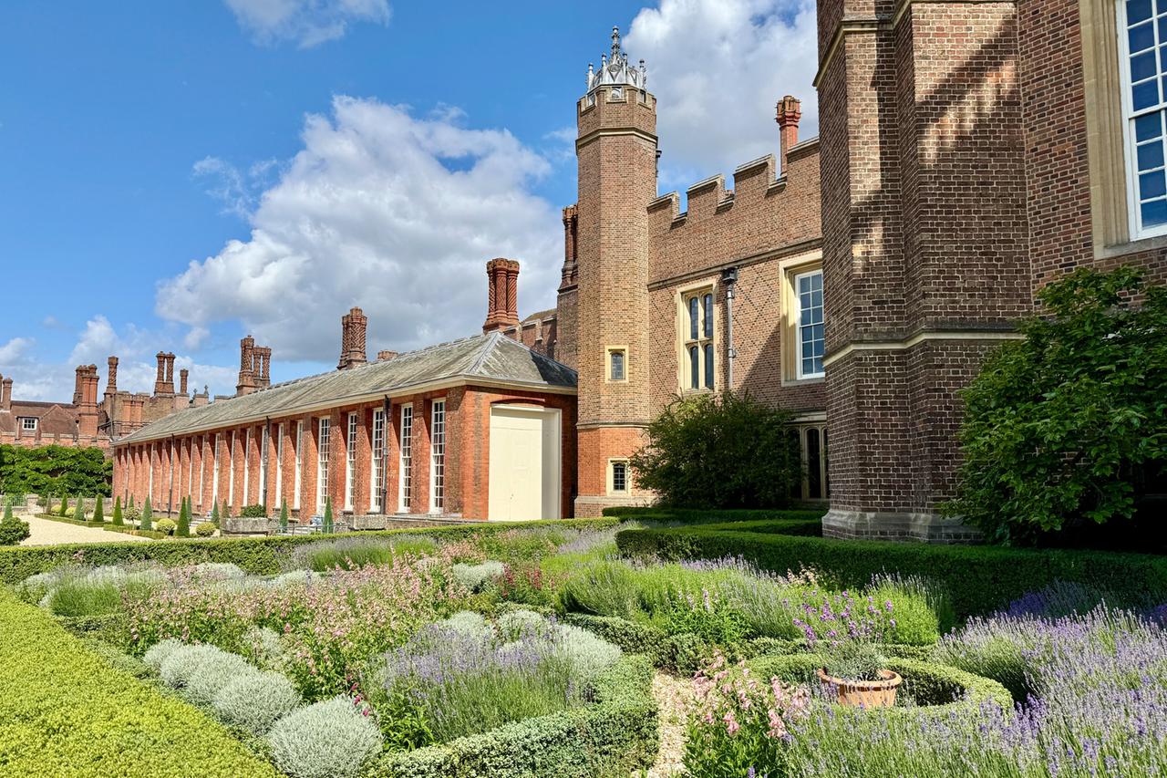 Das Bild zeigt den Knotengarten (Knot Garden) im Hampton Court Palace, einem historischen Palast in England. Im Vordergrund ist der Garten zu sehen, der mit geschwungenen Buchsbaumhecken in geometrischen Formen angelegt ist. Bunte Blumen, darunter einige lila und rosa Blüten, sind in den Beeten gepflanzt.   Im Hintergrund erhebt sich die Backsteinarchitektur des Palastes mit charakteristischen Türmchen und Schornsteinen. Die Fassade zeigt große Fenster, die Tageslicht in die Innenräume lassen. Der Himmel ist blau mit einigen weißen Wolken. Die gesamte Szenerie vermittelt eine harmonische Verbindung von Natur und historischer Architektur sowie ein Gefühl von Ruhe und Ästhetik, das den königlichen Gärten des Palastes eigen ist.
