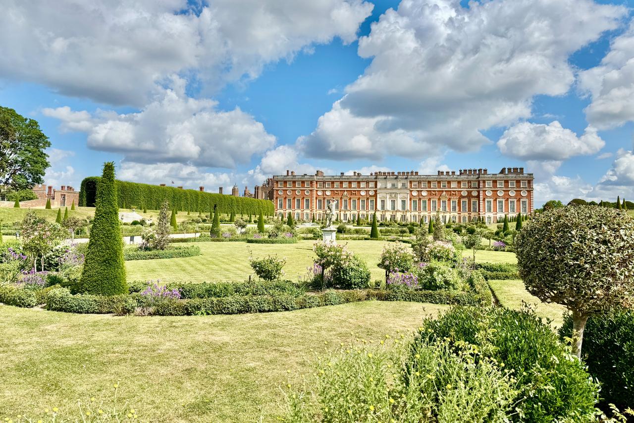 Das Bild zeigt die Südseite des Hampton Court Palace, einem historischen Gebäude, umgeben von gepflegten Gärten. Im Vordergrund erstreckt sich der Privatgarten des Königs, bekannt als der Privy Garden. Die Rasenfläche ist sorgfältig gepflegt und von geometrisch angelegten Beeten mit bunten Blumen und Sträuchern umgeben. Auf der rechten Seite sind mehrere runde und konische Hecken zu sehen.  Der Palace selbst ist in Backsteinbauweise errichtet und hat eine prächtige Fassade mit großen Fenstern und Zierleisten. Das Dach hat eine ansehnliche Anzahl von Schornsteinen, die eine historische Architektur widerspiegeln. Der Himmel ist blau mit einigen weißen, fluffigen Wolken. Die gesamte Szenerie strahlt eine ruhige und harmonische Atmosphäre aus, die das Erbe königlicher Pracht unterstreicht.