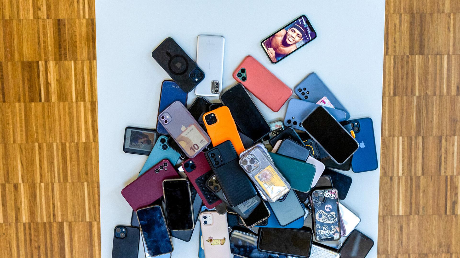 Das Bild zeigt einen Haufen von Mobiltelefonen, die auf einem weißen Tisch abgeladen sind. Die Telefone sind in verschiedenen Farben und Modellen vorhanden, darunter schwarze, graue, blaue, grüne, rote und lilafarbene Geräte. Einige Handys sind ohne Hülle, während andere mit unterschiedlich gestalteten Schutzhüllen versehen sind. Auf einem der Handys ist das Bild eines Menschen zu sehen, möglicherweise ein Selfie. Der Tisch steht auf einem Boden, der aus Holzplanken besteht und eine warme, braune Farbgebung hat. Die Szene wirkt chaotisch und verdeutlicht die große Anzahl an Handys, die hier gesammelt wurden.