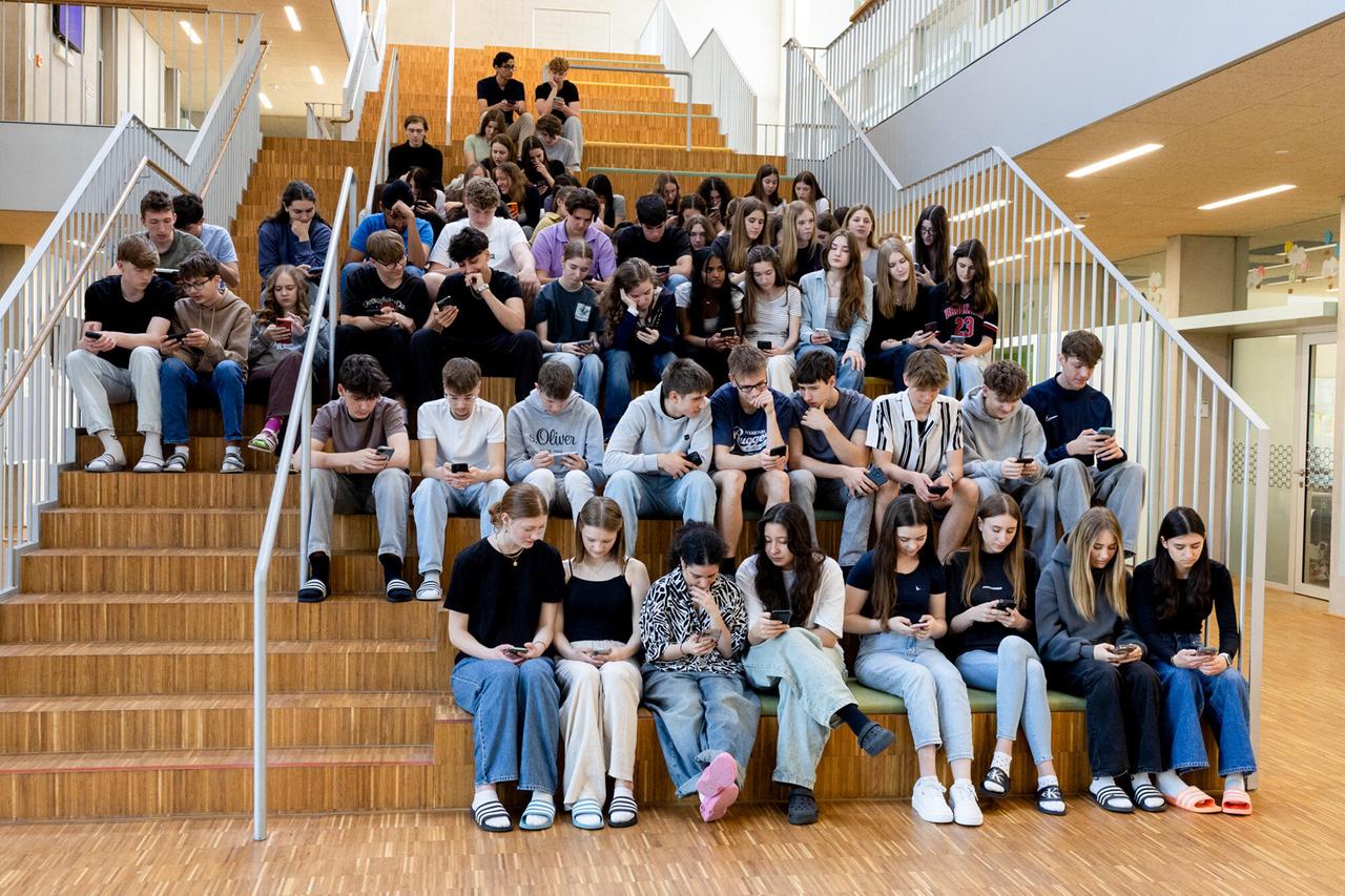 Das Bild zeigt eine Gruppe von Jugendlichen, die auf einer Treppe in einem modernen Raum sitzen. Es gibt insgesamt etwa 40 Jungen und Mädchen, die alle auf ihre Smartphones schauen. Die Treppe hat mehrere Stufen, die aus Holz gefertigt sind, und die Wände sind in neutralen Farben gehalten.   Die Jugendlichen sind unterschiedlich gekleidet, einige tragen T-Shirts, andere Pullover. Ihre Haltungen sind eher entspannt, aber konzentriert, da sie sich mit ihren Geräten beschäftigen. Hinter ihnen sind einige weitere Jugendliche zu sehen, die ebenfalls auf ihren Handys schauen. Der Raum wirkt hell und freundlich, mit großen Fenstern, die viel Licht in den Raum lassen.   Die Situation vermittelt den Eindruck einer Gruppe, die sich in der letzten Minute vor einem Experiment befindet, bei dem sie für drei Wochen auf ihre Handys verzichten müssen.