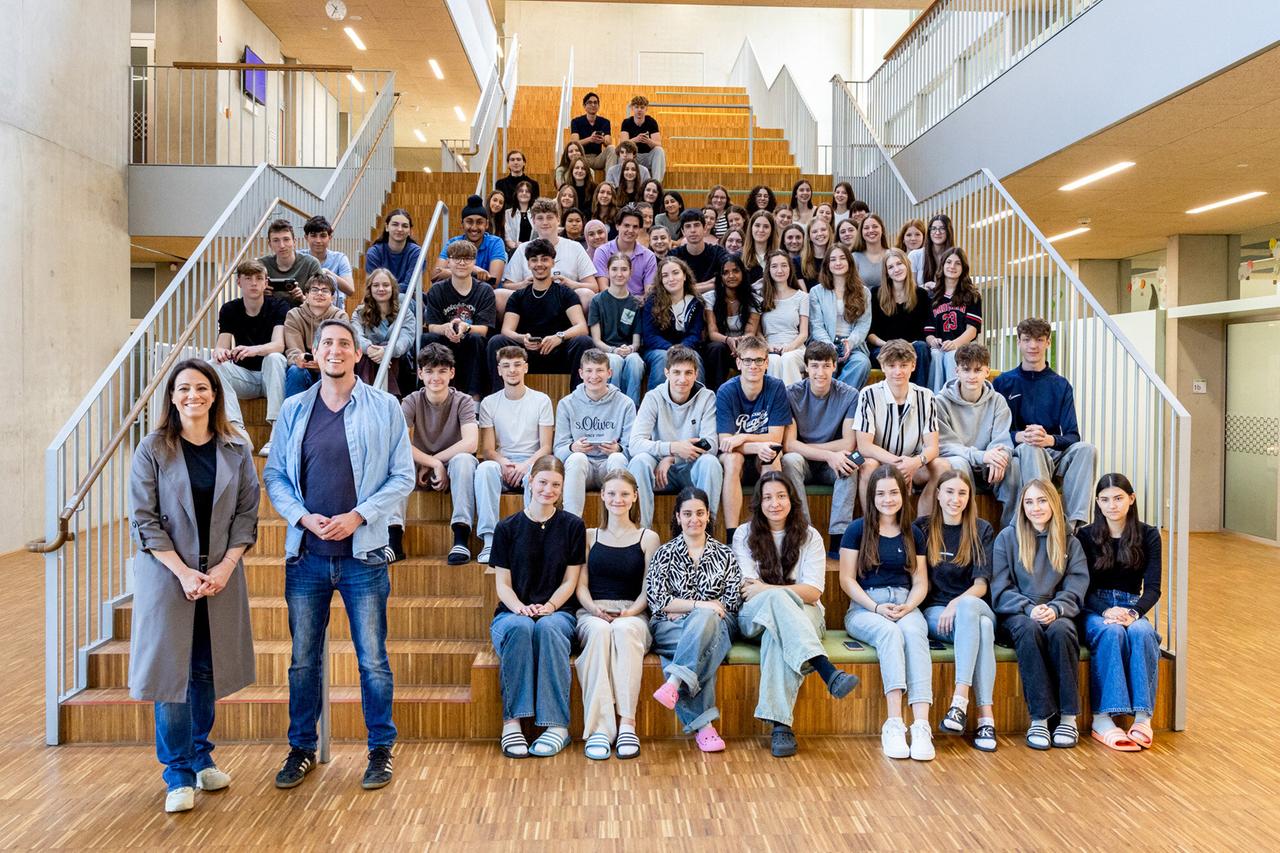 Das Bild zeigt eine Gruppe von Schülern und zwei Lehrpersonen, die auf einer Treppe in einem modernen Gebäude sitzen und stehen. Die Treppe ist aus Holz, und die Wand im Hintergrund ist aus Sichtbeton mit großen Fenstern, die Tageslicht hereinlassen.   In der vorderen Reihe stehen zwei Personen: eine Frau mit langen, dunklen Haaren, die eine graue Jacke trägt, und ein Mann mit kurz geschnittenen, dunklen Haaren, der einen blauen Pullover und eine graue Jacke trägt. Hinter ihnen sitzt eine größere Gruppe von Jugendlichen, die in verschiedenen Freizeitkleidungen sind. Es sind sowohl Jungen als auch Mädchen vertreten, und viele tragen lässige T-Shirts und Jeans.  Die Gesichtsmimik der Schüler ist überwiegend neutral bis freundlich, und die Szene strahlt eine Atmosphäre von Zusammenhalt und Gruppendynamik aus. Insgesamt sind etwa 69 Jugendliche im Bild, vor einer Kulisse, die modern und einladend wirkt.