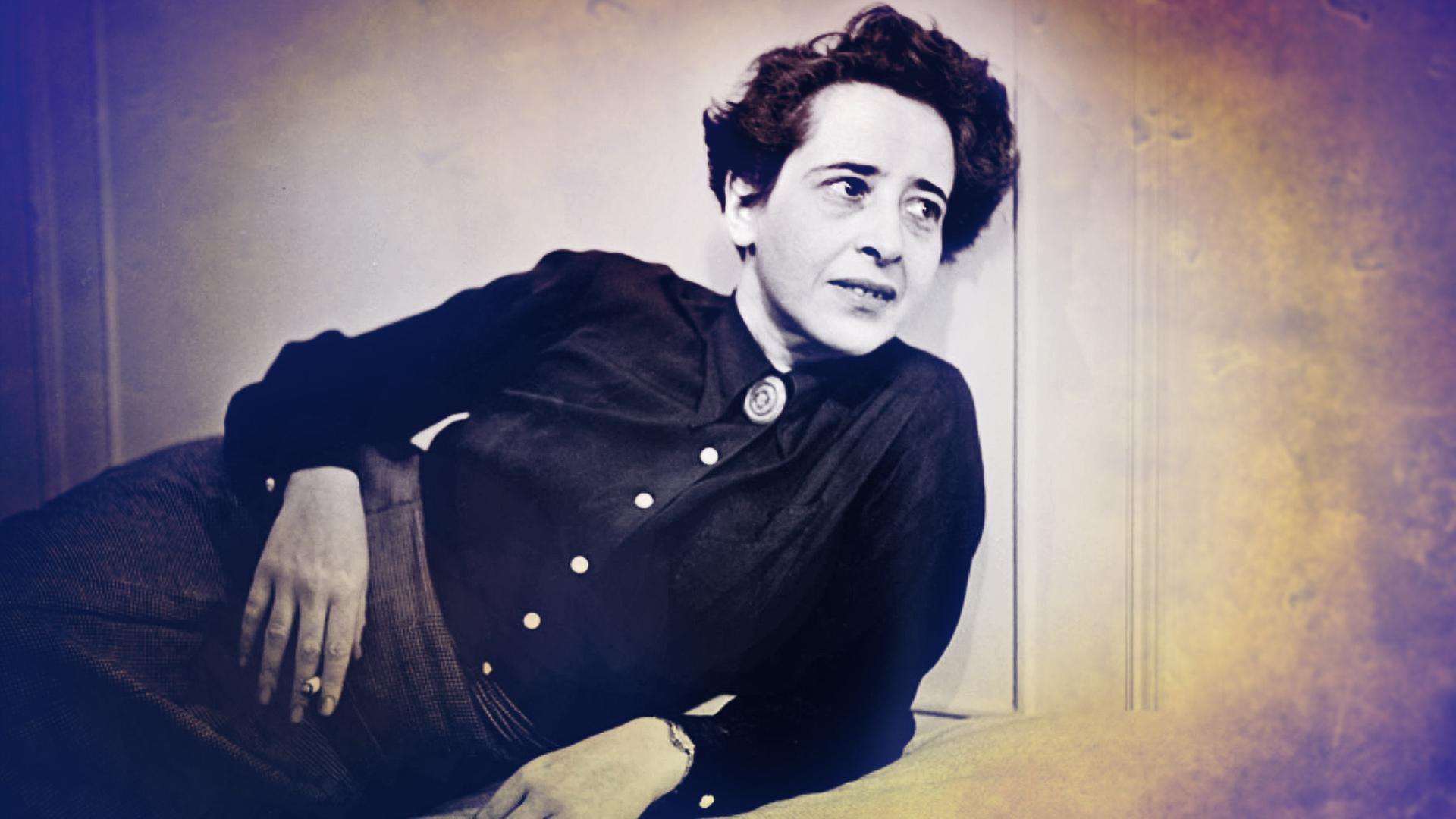 Hannah Arendt sitzt auf einem Sessel bei einem Fernsehinterview. Sie trägt ein schwarzes Kostüm und ihr Ellenbogen stützt sie auf der Armlehne auf. IN der Hand hält sie eine ZIgarette. Das Bild ist schwarz weiß. 