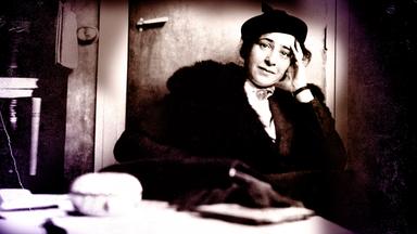 Hannah Arendt sitzt auf einem Sessel bei einem Fernsehinterview. Sie trägt ein schwarzes Kostüm und ihr Ellenbogen stützt sie auf der Armlehne auf. IN der Hand hält sie eine ZIgarette. Das Bild ist schwarz weiß. 