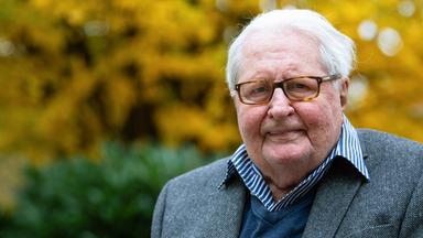 Ein alter Mann mit Brille und grauem Jacket vor einem verschwommenen Hintergrund.