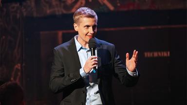 Till Reiners steht in der Mitte des Bildes und hebt seine linke Hand. In der rechten Hand hält er ein Mikrofon. Till Reiners trägt ein dunkles Sakko mit hellblauem Hemd. Der Hintergrund ist dunkel.