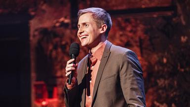 Moderator steht in der Mitte des Bildes und blickt an den linken Bildrand. In seiner rechten Hand hält er ein Mikrofon. 