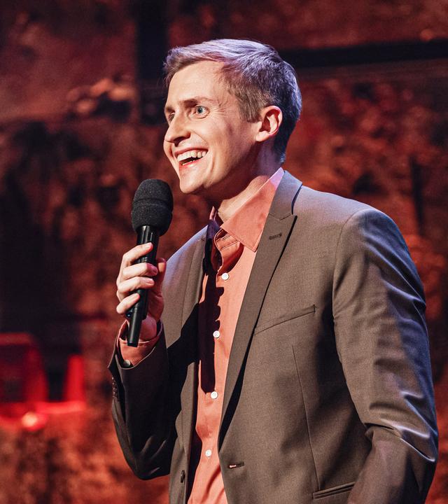 Moderator steht in der Mitte des Bildes und blickt an den linken Bildrand. In seiner rechten Hand hält er ein Mikrofon. 