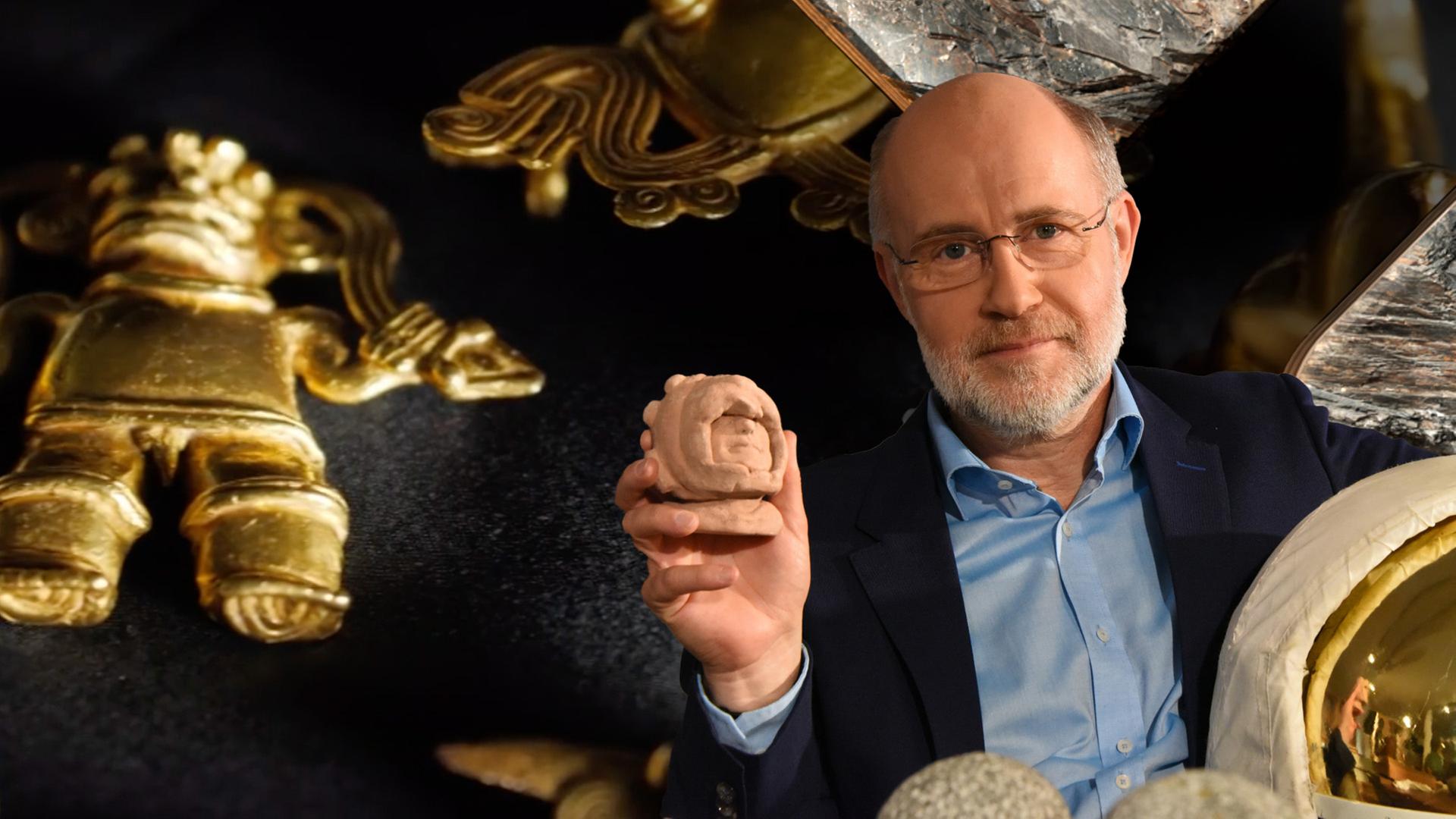 Harald Schmitt hält ein Artefakt hoch: ein Teil einer Lehmfigur eines menschliches Gesichts.