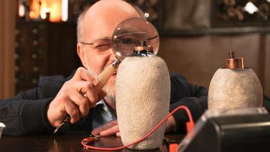 Harald Lesch betrachtet durch eine große Lupe zwei Nachbauten einer "Bagdad-Batterie".