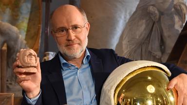 Harald Lesch hält in der einen Hand ein kleine Mayaskulptur und in der anderen eine Art Taucherhelm.