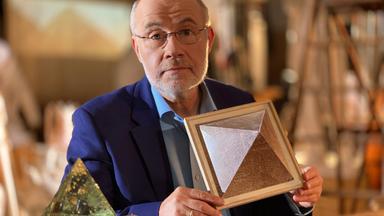 Moderator Harald Lesch sitzt an einem Tisch und hält das Modell einer Pyramide in seinen Händen.