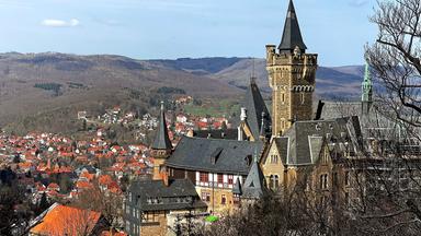 Ein Schloss thront über einer Ansiedlung von Häusern. Im Hintergrund sind Hügel zu sehen.