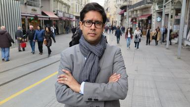 Ein Mann mit Brille, Schal und Jacket steht mit verschränkten Armen inmitten einer Fußgängerzone.