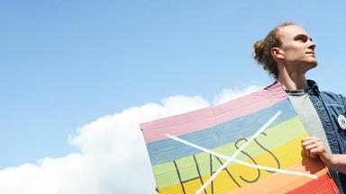 Florian hält ein buntes Schild vor seinen Körper. Auf dem Schild ist das Wort "Hass" durchgestrichen. Er blickt zur Seite.