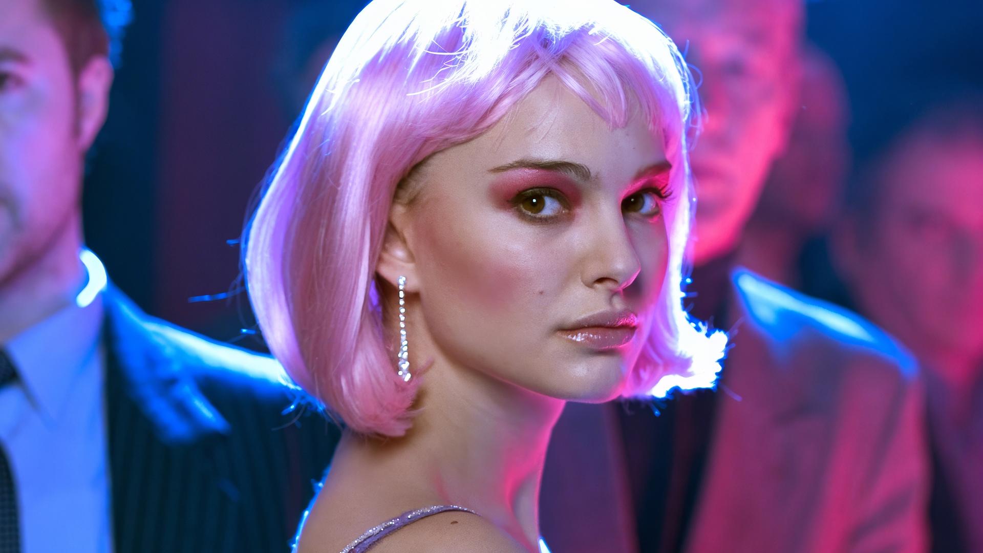 Alice (Natalie Portman) trägt eine pinkfarbene Perücke, auffälliges Make-up und blickt ernst in die Kamera. Sie steht inmitten einer Gruppe von Menschen in einem in blaues und pinkes Licht getauchten Raum.