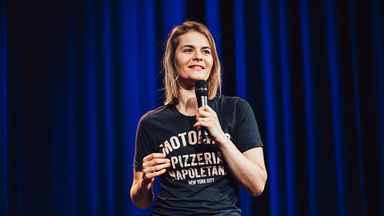 Hazel Brugger steht auf der Bühne vor einem blauen Vorhang.