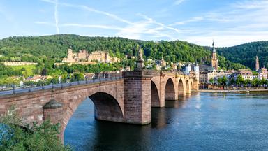 Eine alte Brücke führt über den Neckar. Am anderen Ufer sind eine Schlossruine und die Altstadt von Heidelberg mit einer Kirche zu sehen.