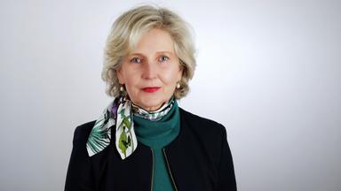 Porträtfoto einer Frau mittleren Alters mit kurzen graumelilerten Haaren, die ein schwarze Jacke über einem Rollkragenpullover und kurzen Schal trägt.