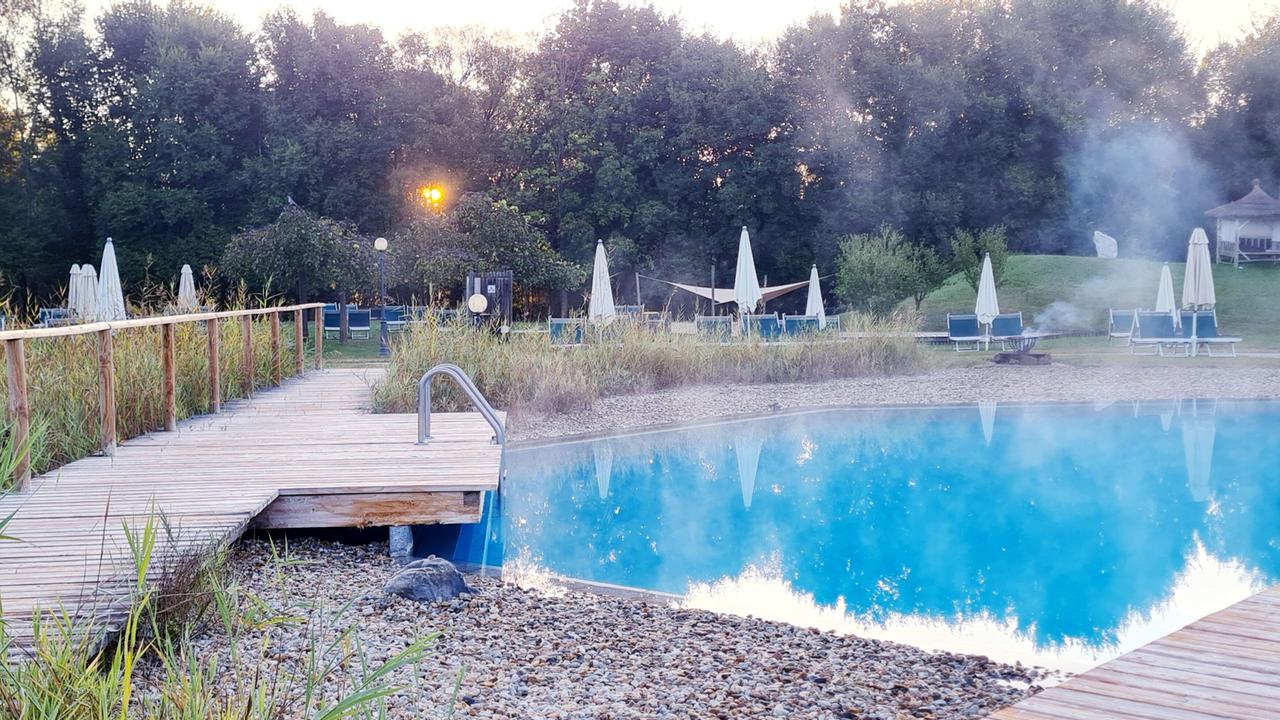 Das Bild zeigt einen ruhigen Spa-Bereich in der Natur, insbesondere die Therme Blumau - Vulkania. Im Vordergrund führt ein hölzerner Steg an einen blauen Thermal-Wasserpool, dessen Oberfläche leicht dampft. Um den Pool herum sind einige Steine und pflanzliche Elemente wie Gras und Sträucher.   Im Hintergrund sind mehrere Liegen mit weißen Sonnenschirmen angeordnet, umgeben von Bäumen und einer ruhigen Umgebung. Die Sonne steht tief am Horizont und erzeugt eine friedliche Atmosphäre. Neben den Liegen sind auch einige Bäume und eine kleine Grünfläche sichtbar. Es scheint ein ruhiger und entspannender Ort zu sein, der für Wellness und Erholung genutzt wird.