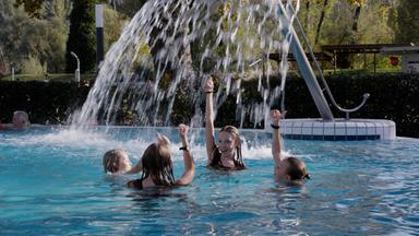 Das Bild zeigt eine fröhliche Szene in einem Außenpool, wahrscheinlich in einem Thermalbad. Im Vordergrund sind vier Kinder zu sehen, die im Wasser spielen. Sie haben nasse Haare und tragen Badebekleidung. Die Kinder zeigen mit ihren Händen nach oben, offenbar erfreut über das Wasser, das aus einer etwa zwei Meter hohen Wasserrampe herabfließt.  Im Hintergrund ist ein älterer Mann zu erkennen, der entspannt im Wasser sitzt. Der Pool ist von einer grünen Hecke umgeben, und im Hintergrund sind Bäume und weitere Freizeitbereiche sichtbar. Der Himmel scheint sonnig, was auf angenehmes Wetter hindeutet. Die gesamte Szene vermittelt ein Gefühl von Freude, Erholung und Freizeitaktivität.