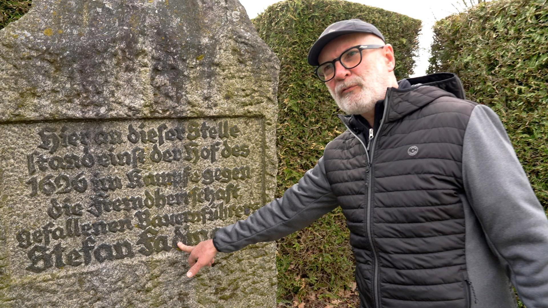 Ein Mann zeigt auf einen Namen auf einem verwitterten Gedenkstein