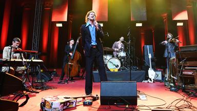 Ein Mann auf der Bühne singt in ein Mikrofon. Andere Musiker im Hintergrund.