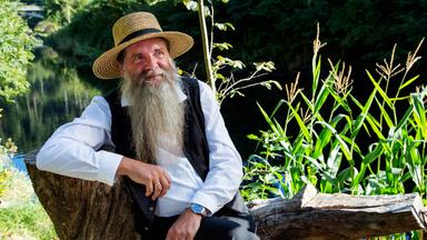 Ein Mann mit langem Bart und Strohhut sitzt auf einem Baumstamm im Freien.