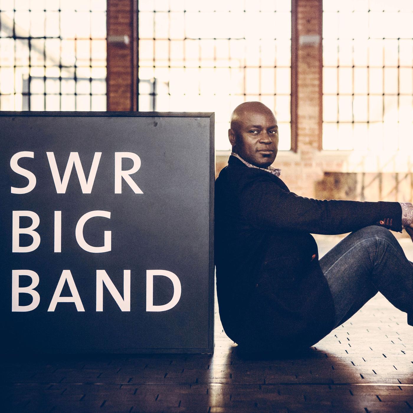 Der Sänger Ola Onabulé sitzt auf dem Boden und lehnt an einem Schild, auf dem SWR BIG BAND steht.