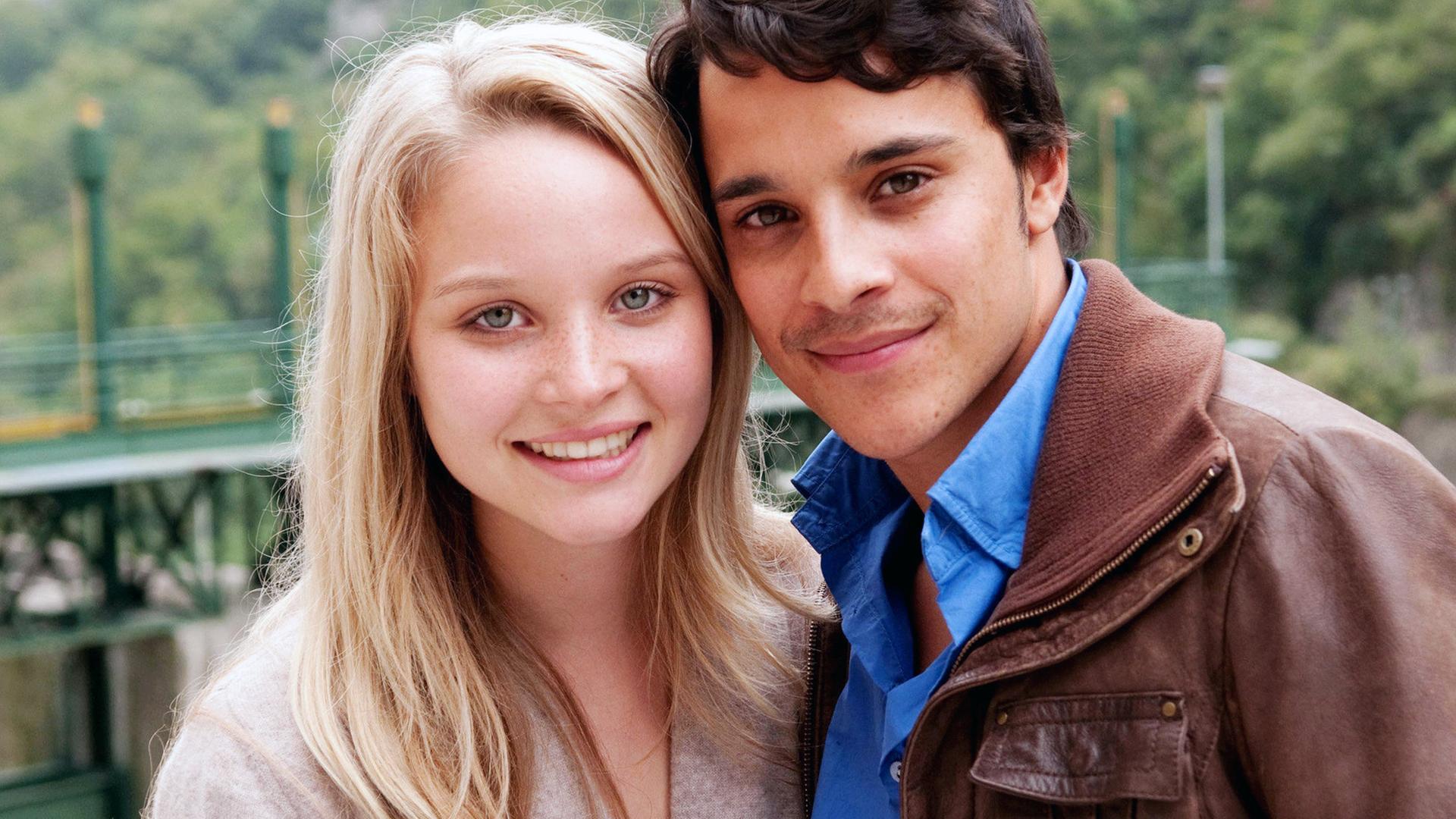 Eine blonde, junge Frau und ein dunkelhaariger, junger Mann stehen seitlich aneinander gelehnt und schauen glücklich in die Kamera