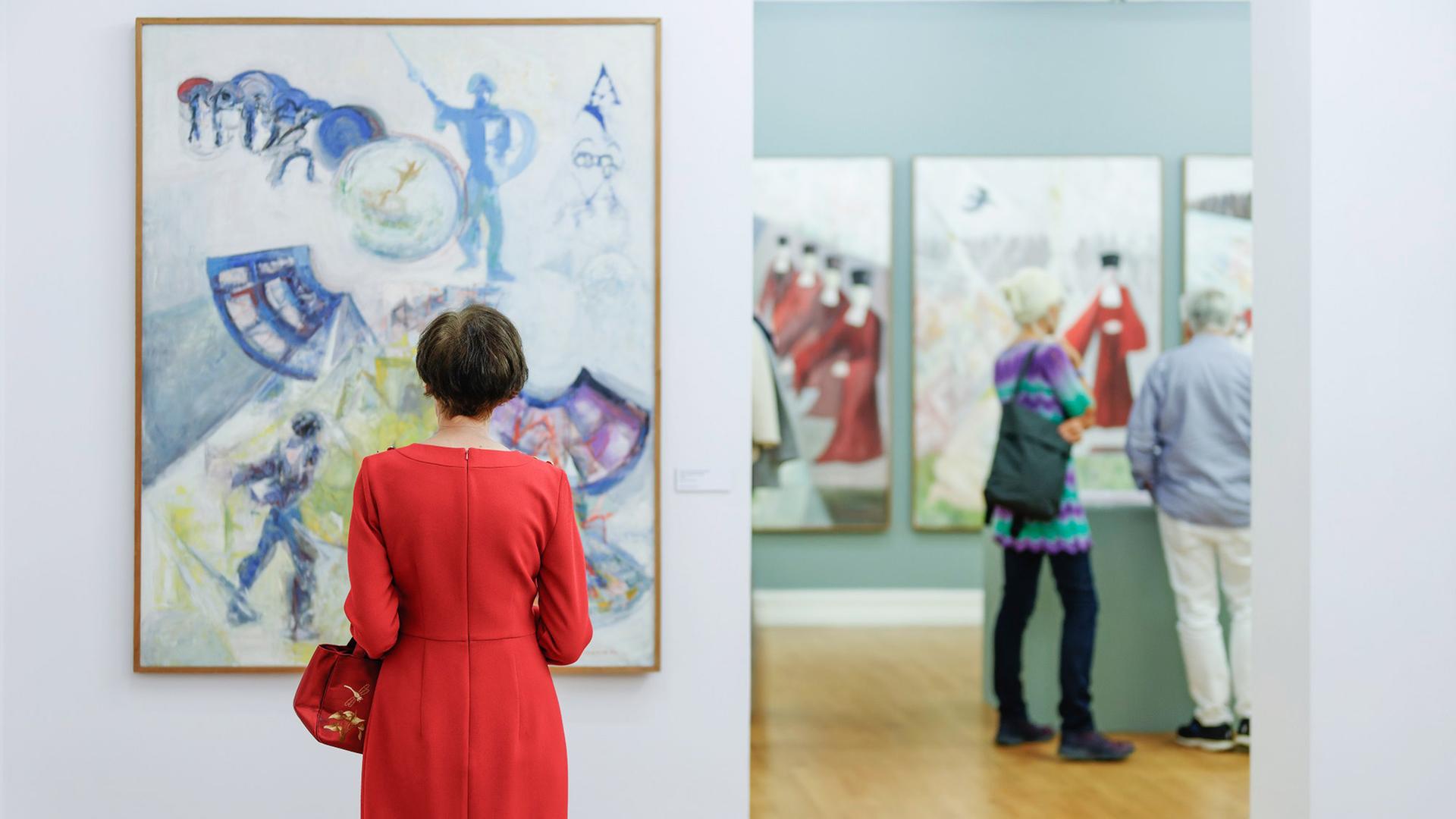 Rückansicht einer Frau im roten Kleid, die ein großformatiges Gemälde aus vielen pastellfarbenen Strichen und Formen betrachtet. Im Hintergrund stehen weitere Personen vor Gemälden.