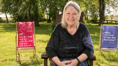 Eine Frau mit grauen Haaren sitzt im Freien und lächelt.
