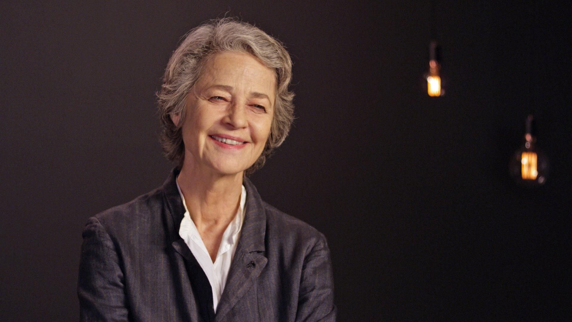 Auf dem Bild sieht man die Schauspielerin Charlotte Rampling in einer Interviewsituation vor sehr dunklem Hintergrund. Sie trägt ein dunkles Oberteil mit weißem Kragen, kurzes graues Haar und lächelt dem Interviewpartner zu.