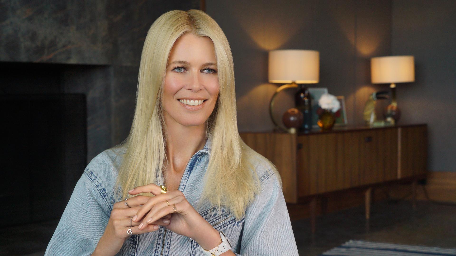 Man sieht das ehemalige Model Claudia Schiffer sitzend in einer Interviewsituation. Sie trägt ein Jeans-Oberteil und ihr langes blondes Haar offen. Im Hintergrund stehen zwei Tischlampen und ein Sideboard.