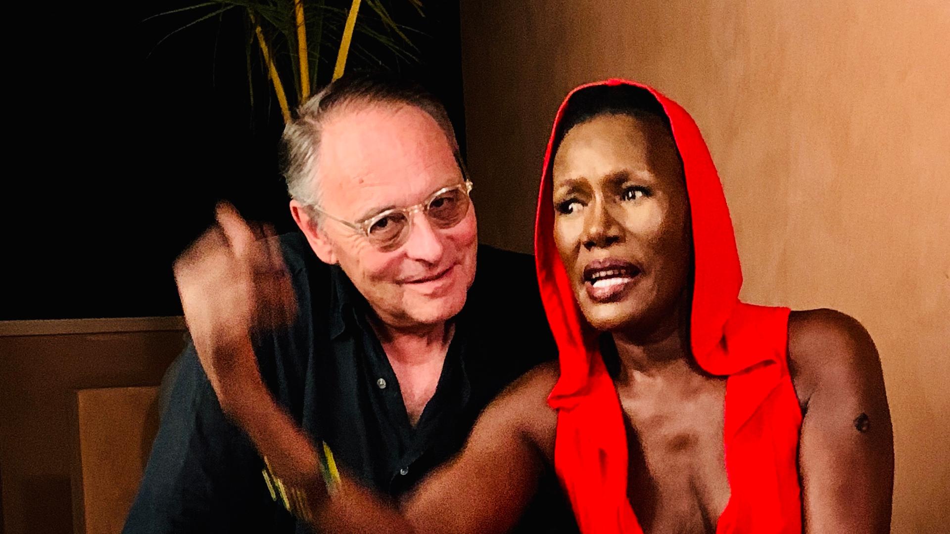 Auf dem Foto sieht man die Schauspielerin Grace Jones und den Filmemacher Gero von Boehm. Die beiden sitzen nebeneinander. Die Frau trägt einen tief ausgeschnittenen, roten Overall mit Kapuze und ist barfuß, ein Bein liegt auf der Matratze, auf der sie sitzt. Der Mann trägt ein dunkles Hemd, helle Hose und Brille und lächelt in die Kamera.