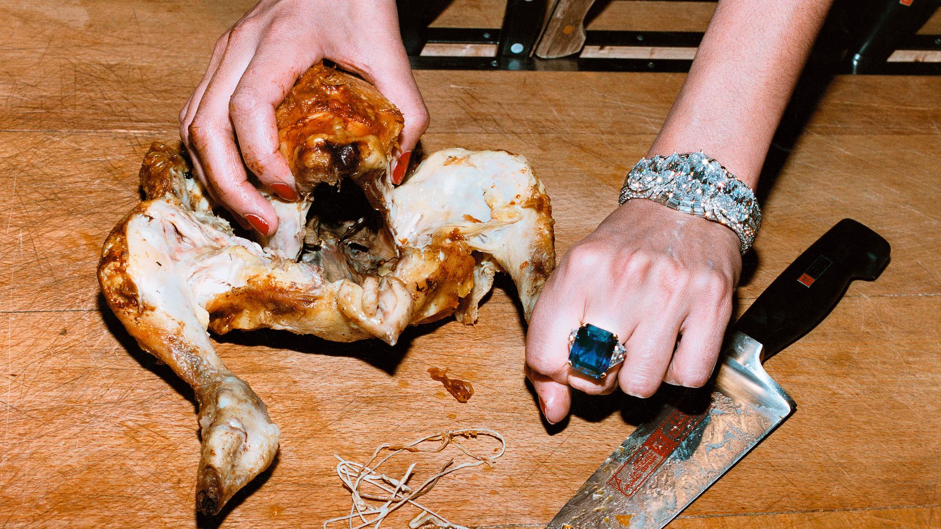 Auf dem Bild sieht man die Hände einer Frau, die gerade eine Brathühnchen auf einer Holzplatte präpariert. An ihrer linken Hand trägt sie einen großen blauen Edelsteinring und ein großes, silbernes Juwelenarmband.