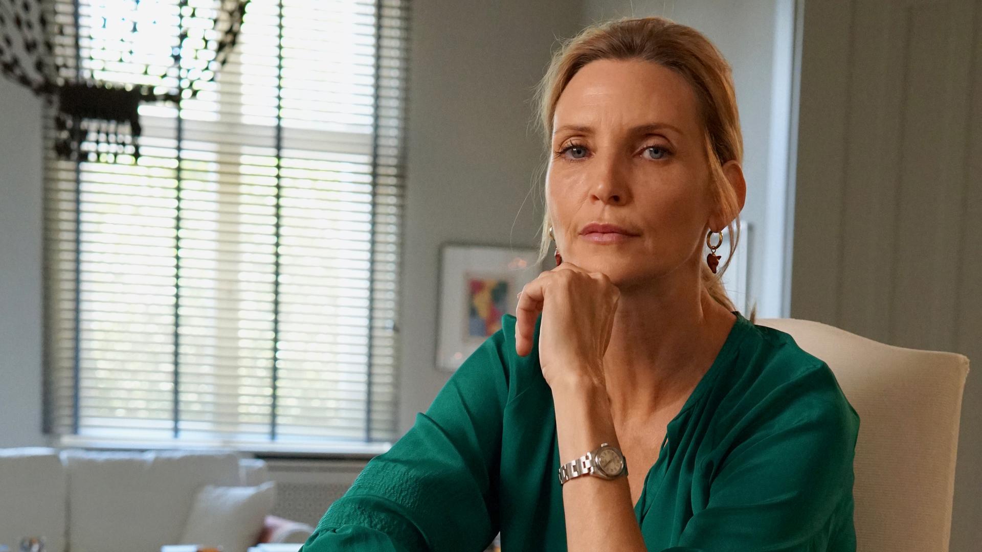 Auf dem Bild sieht man das Model Nadja Auermann. Sie sitzt auf einem Stuhl in einem großen Zimmer und hat die Beine übereinander geschlagen. Den Arm stützt sie auf und legt das Kinn auf die Hand. Sie schaut nachdenklich. Sie trägt eine schwarze Hose und eine dunkelgrüne Bluse.