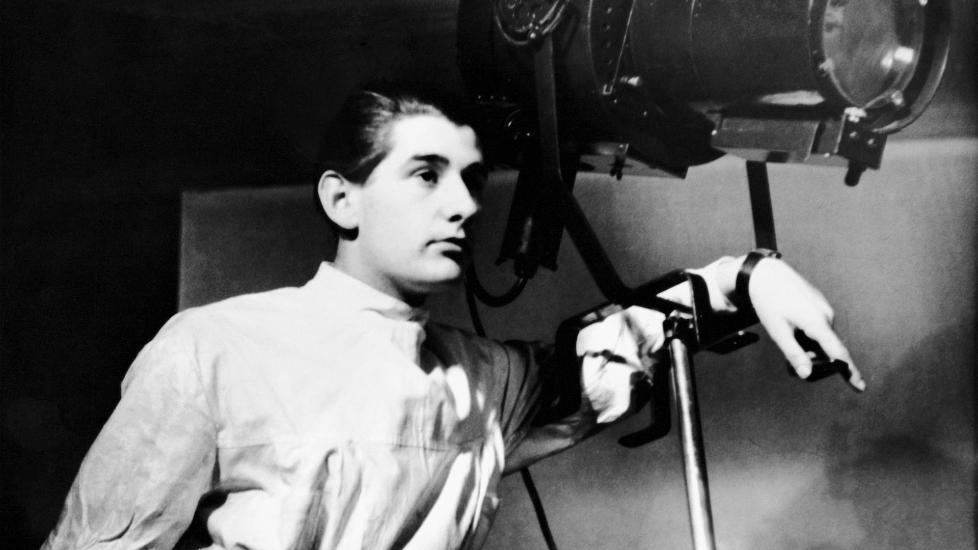 Man sieht ein schwarz-weiß Foto des sehr jungen Helmut Newton in einem weißen Kittel. Er lehnt an einen Fotoscheinwerfer und schaut lässig zur rechten Bildkante.