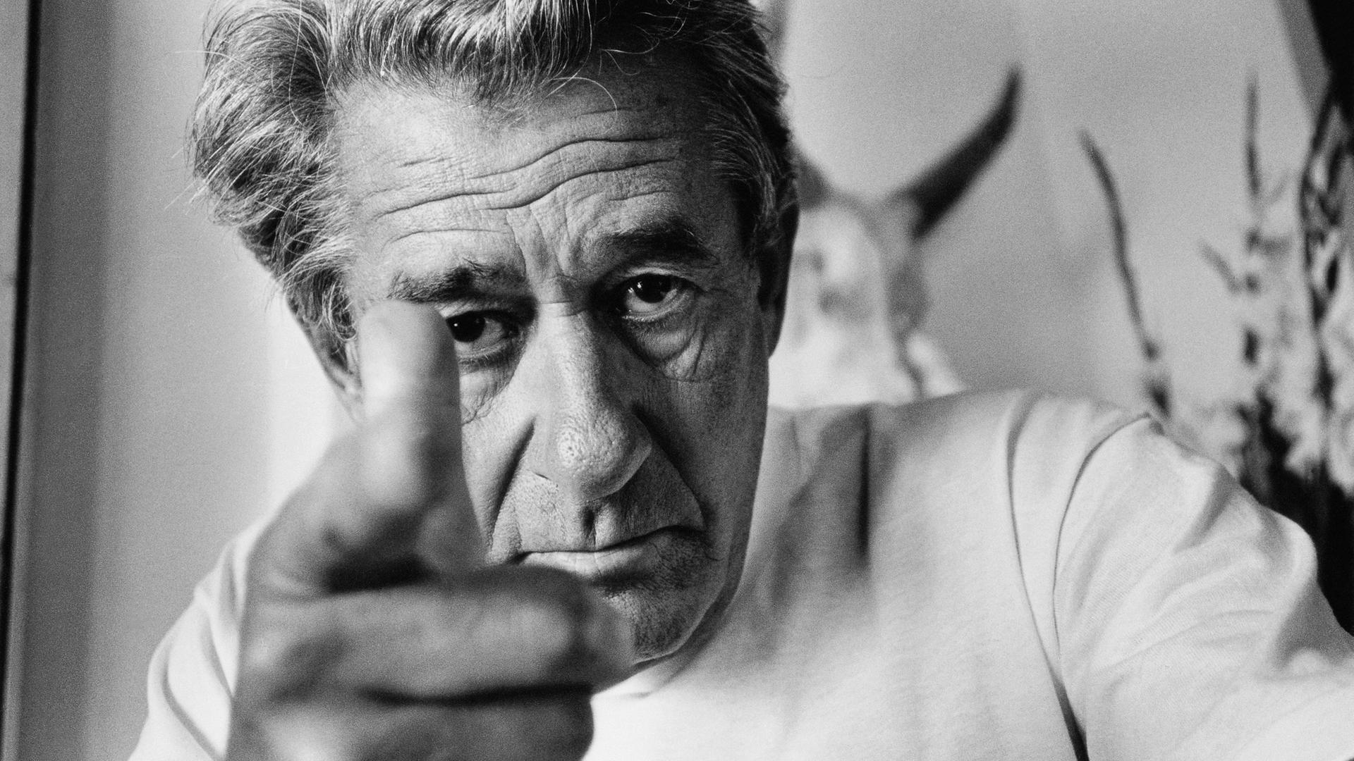 Auf dem schwarz-weiß Bild sieht man den Fotografen Helmut Newton,  er schaut direkt in die Kamera und zeigt mit seinem rechten Zeigefinger direkt auf den Betrachter.