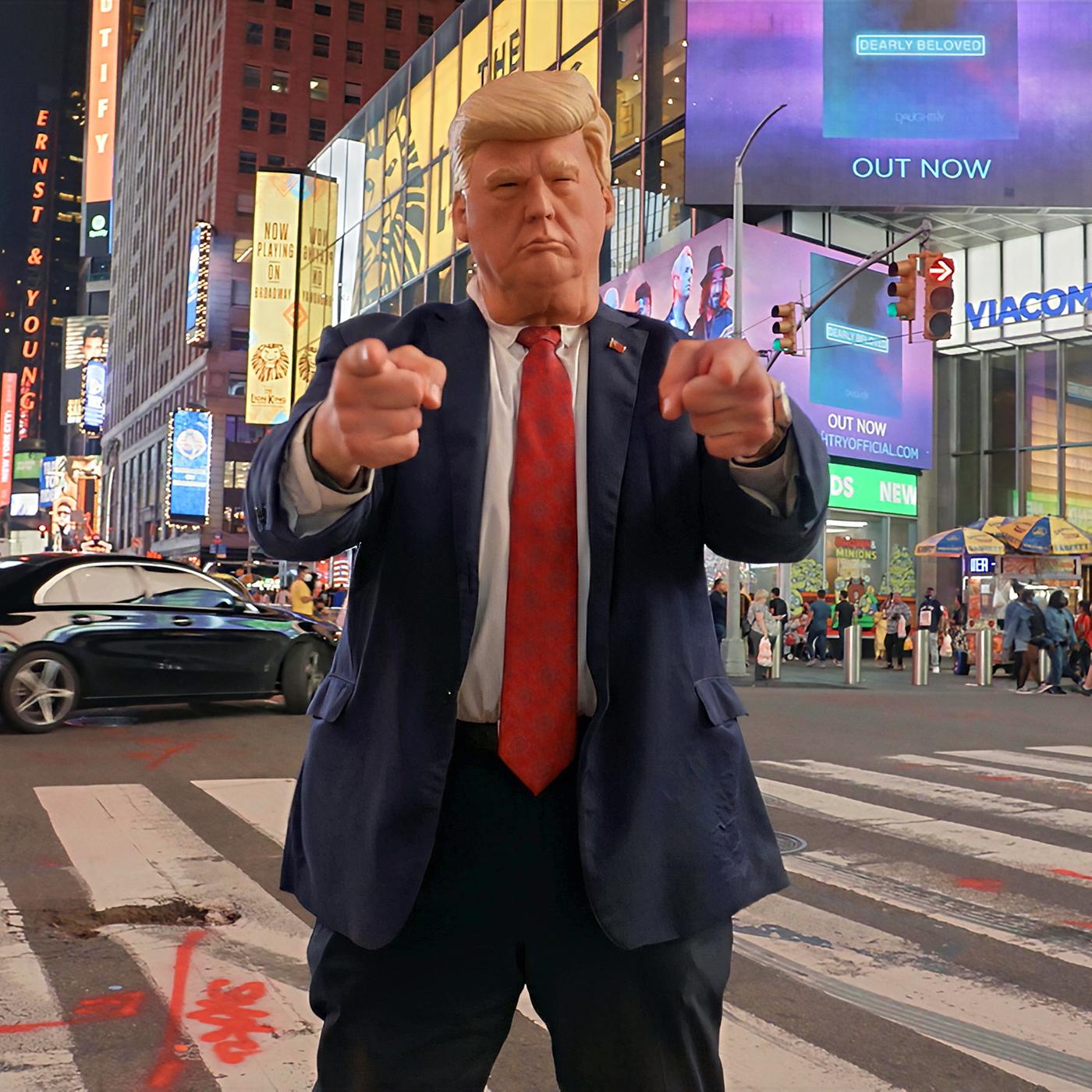 Ein Donald-Trump-Impersonator mit Maske steht nachts am Times Square in New York auf einem Zebrastreifen und zeigt mit beiden Händen in die Kamera.