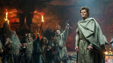 Elrond steht in einem dunklen Raum, der mit Fackeln ausgeleuchtet ist. Im Hintergrund sind mehrere Zwerge zu erkennen, die gestikulieren.