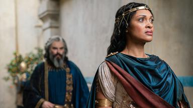 Míriel steht vor einem Gebäude. Sie wirkt skeptisch. Pharazôn ist im Hintergrund in der Unschärfe zu sehen.