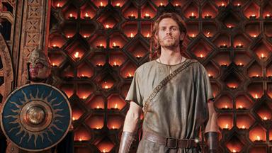 Halbrand steht vor einer Wand, die mit brennenden Kerzen beleuchtet ist. Neben ihm stehen zwei Wachen (Komparsen).