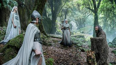 Der Fremde steht in einem Wald den drei Mystikerinnen gegenüber. Die Frauen sind mit hellen Gewändern bekleidet. Eine von ihnen trägt einen Helm, die anderen beiden Kopftücher.