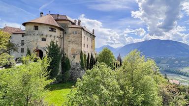 Schloss Schenna thront auf einem Hügel über Meran, umgeben von Bäumen und mit Blick auf die Berglandschaft Südtirols.