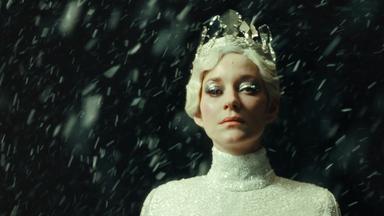 Porträtfoto einer weißblonden Frau mit silbrig glitzerndem Diadem in einem engen silbgrig glitzernden Rollkragen-Oberteil mit dickem silberfarbenem Lidschatten vor einem dunklen Hintergrund.